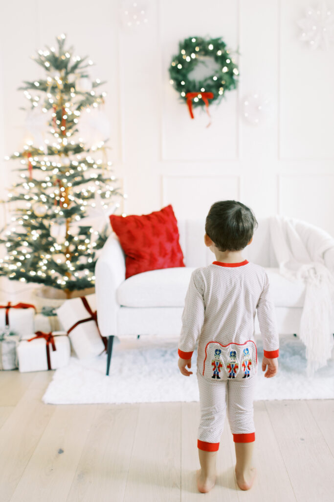 little boy in christmas jammies in front of white couch in a studio thats decorated for christmas  with a wreath and christmas tree in the background