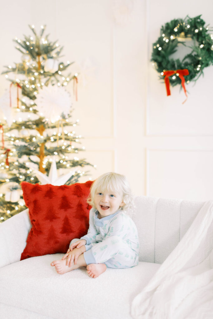 toddler boy in christmas jammies sitting on a white couch in a studio thats decorated for christmas  with a wreath and christmas tree in the background