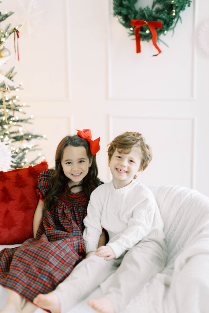 siblings wearing their chritmas dresses sitting on a white couch in a studio thats decorated for christmas  with a wreath and christmas tree in the background