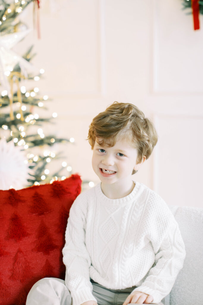 preschool aged boy in a white sweater sitting on a couch in a studio decorated for chritmas 