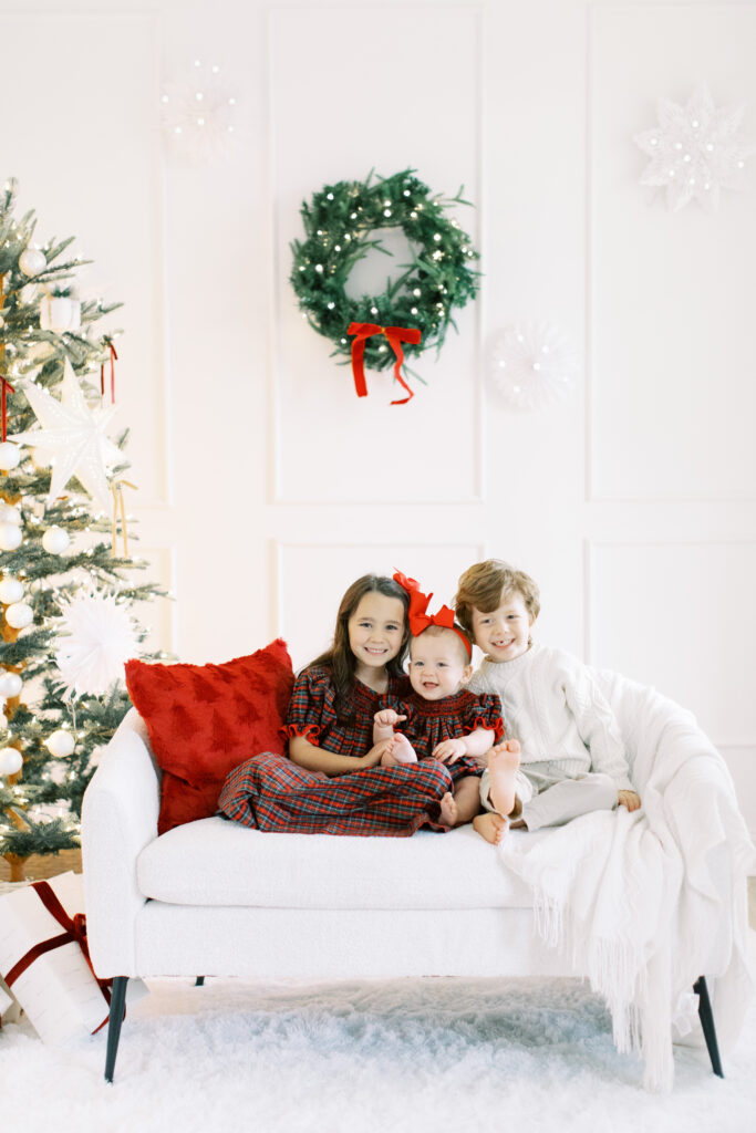 siblings wearing their chritmas dresses sitting on a white couch in a studio thats decorated for christmas  with a wreath and christmas tree in the background